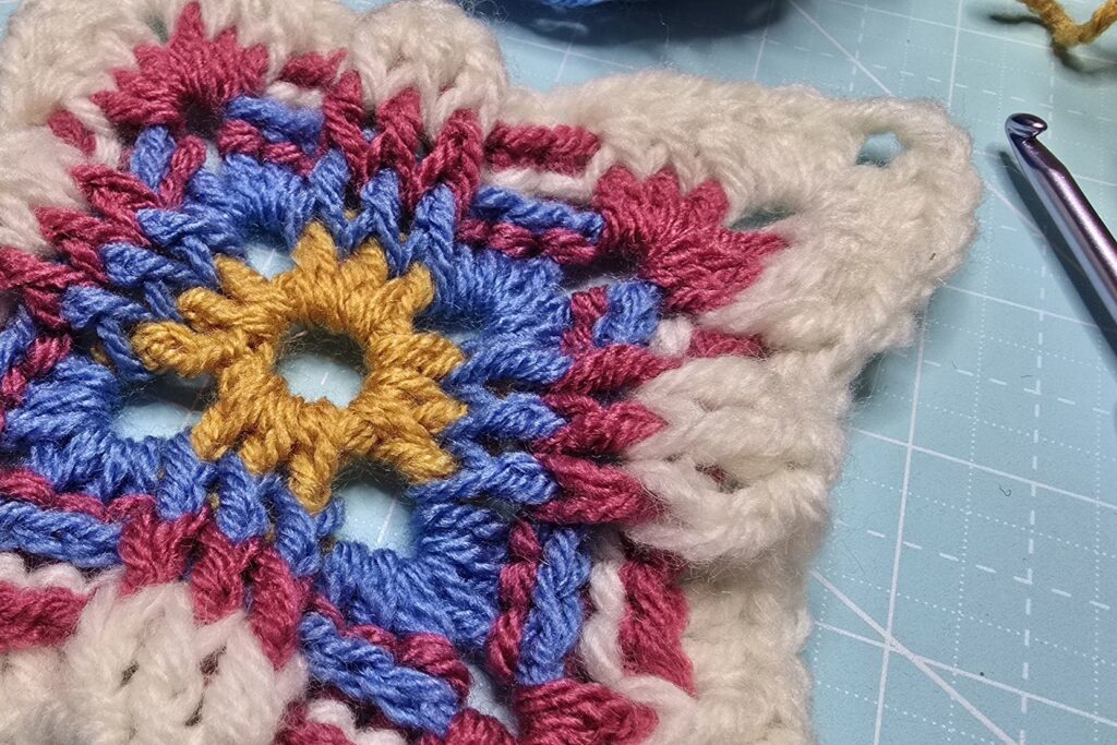 A closer Russian granny square with combinations of gold, blue, magenta, and white yarns showcasing how the front and back post double makes the square looks stunning